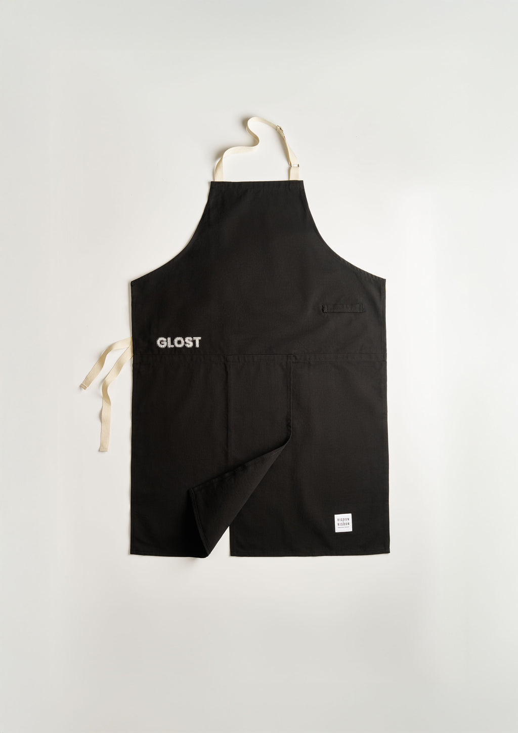 The GLOST potter's apron laid flat: black cotton with GLOST embroidered in white, cream cotton ties, a chest pocket, and the split skirt folded back to show the opening.