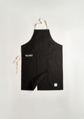 The GLOST potter's apron laid flat: black cotton with GLOST embroidered in white, cream cotton ties, a chest pocket, and the split skirt folded back to show the opening.