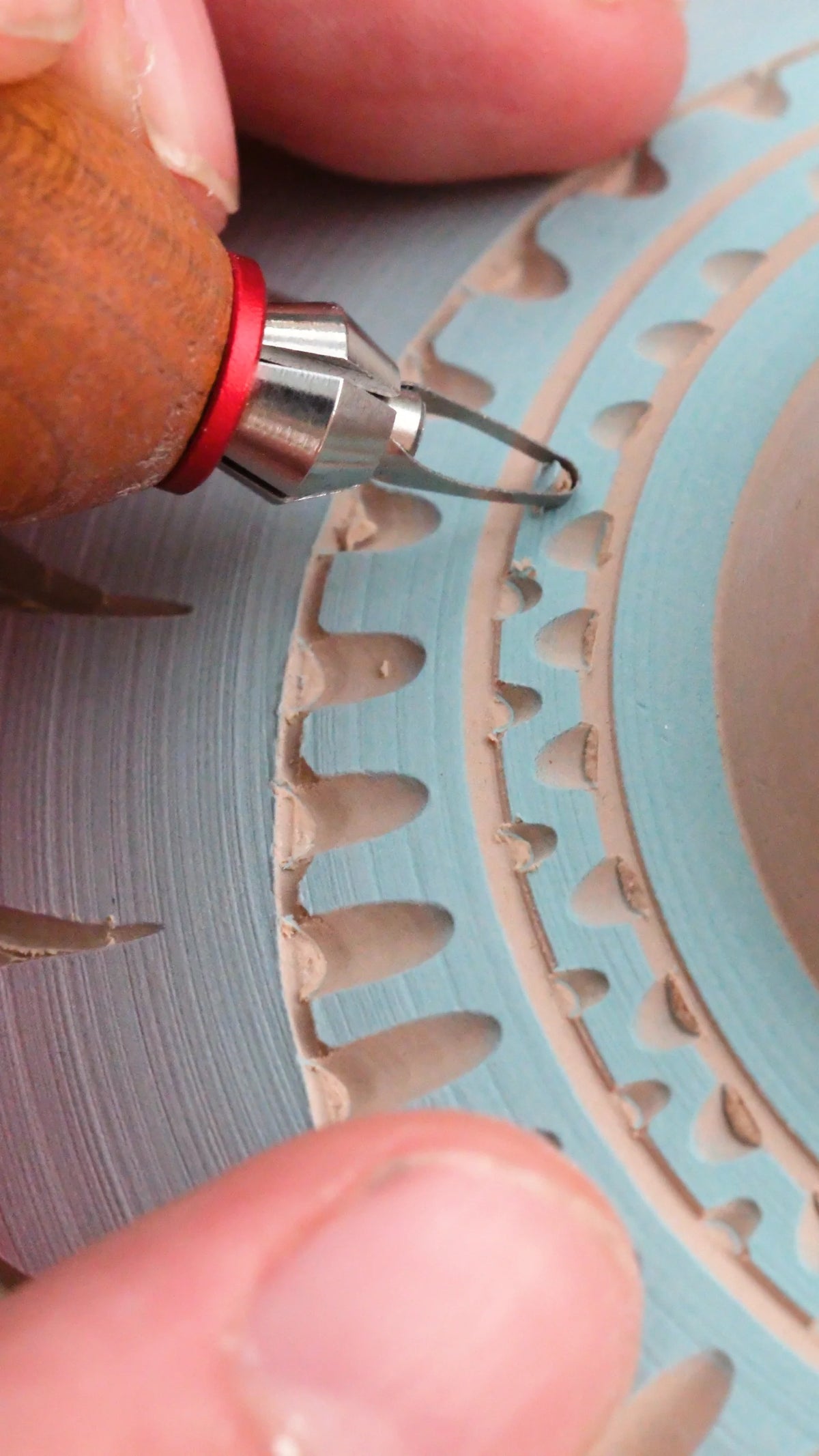 A BuschKraft Dragonfly blade carving a decorative border into a plate covered in blue slip