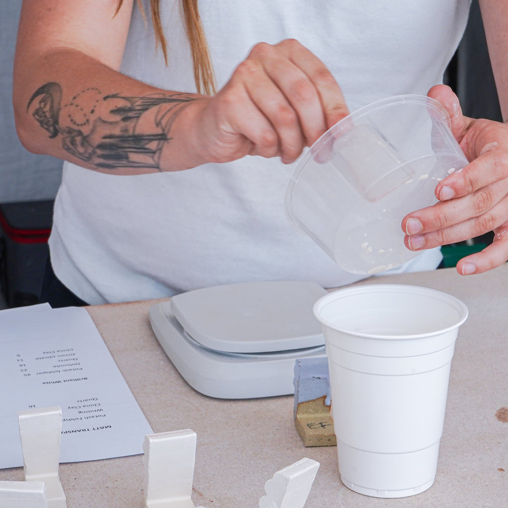 On the table, there are white ceramic test tiles, a scale, and a sheet of paper with notes, suggesting a glaze mixing or testing process. Various materials and tools are organised in the background, enhancing the hands-on, instructional nature of the setting.