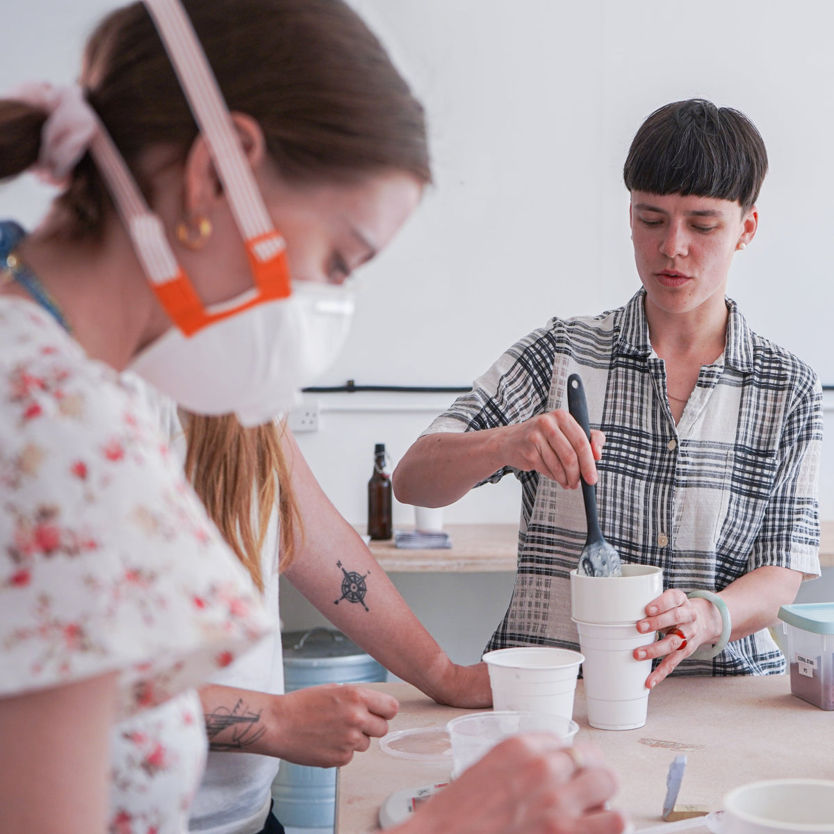 Three individuals are working together in a ceramic studio. The person in the foreground, wearing a floral shirt and a protective mask, is focused on mixing materials. Another individual, in a checkered shirt, is stirring glaze or clay in a cup, demonstrating the process. Various containers and tools are scattered on the table, and a kiln can be seen in the background. The atmosphere suggests a hands-on workshop involving ceramic glaze preparation.