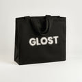 The GLOST cotton tote standing upright, black canvas with the GLOST name printed in white dotted type and black webbing handles folded down.