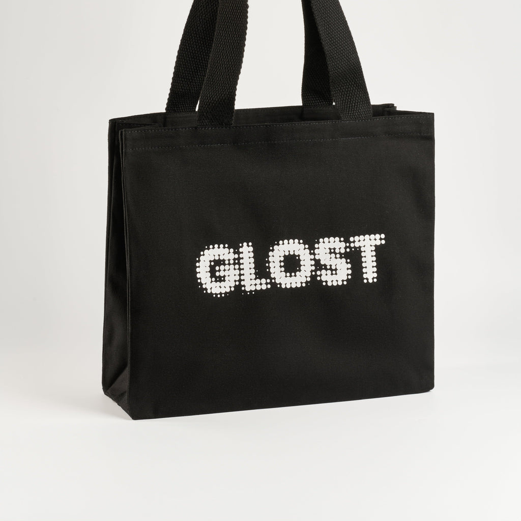 The GLOST black cotton tote seen from the side with its webbing handles raised.