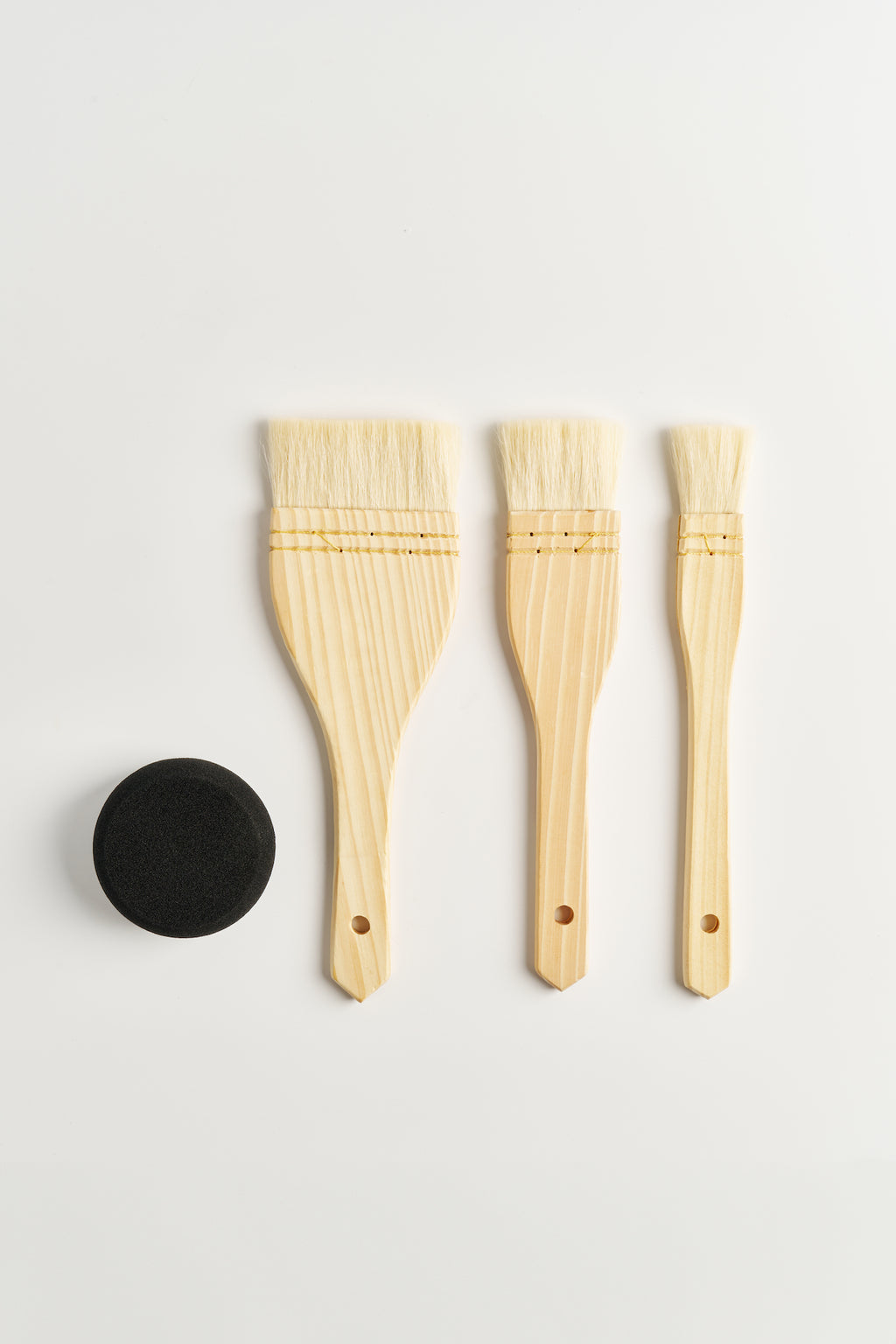 Three hake brushes laid in a row with a round black sponge, seen from above
