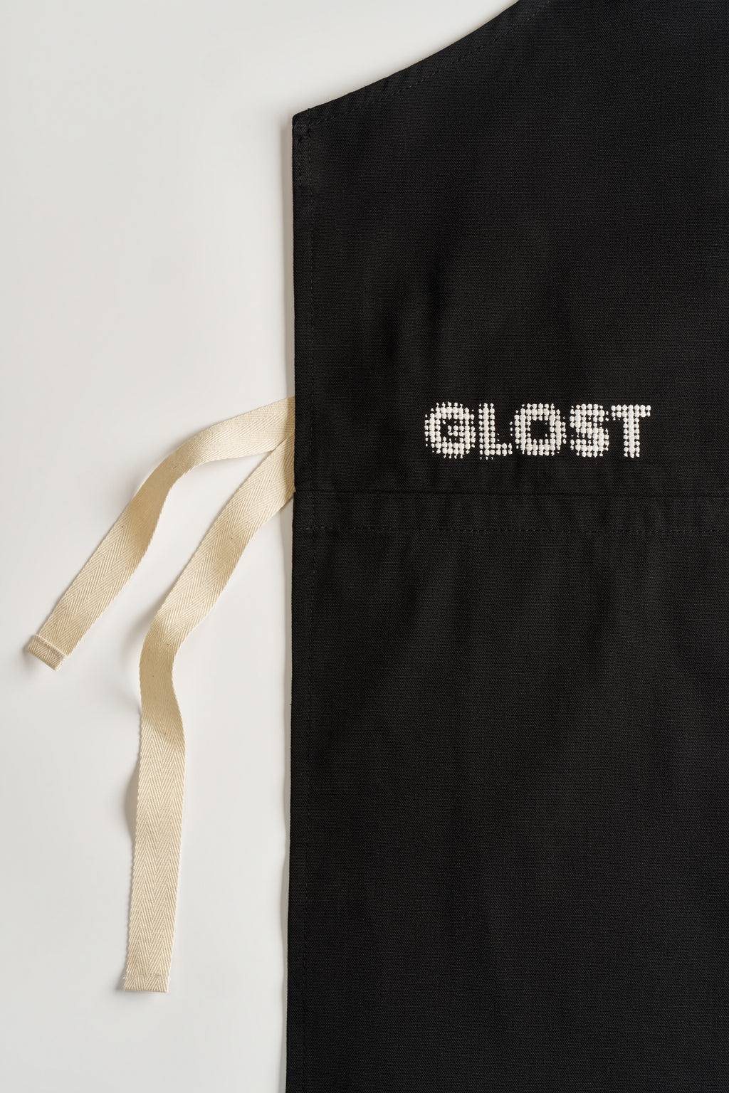 Close view of the GLOST name embroidered in white on the black cotton apron, with the cream cotton waist ties alongside.