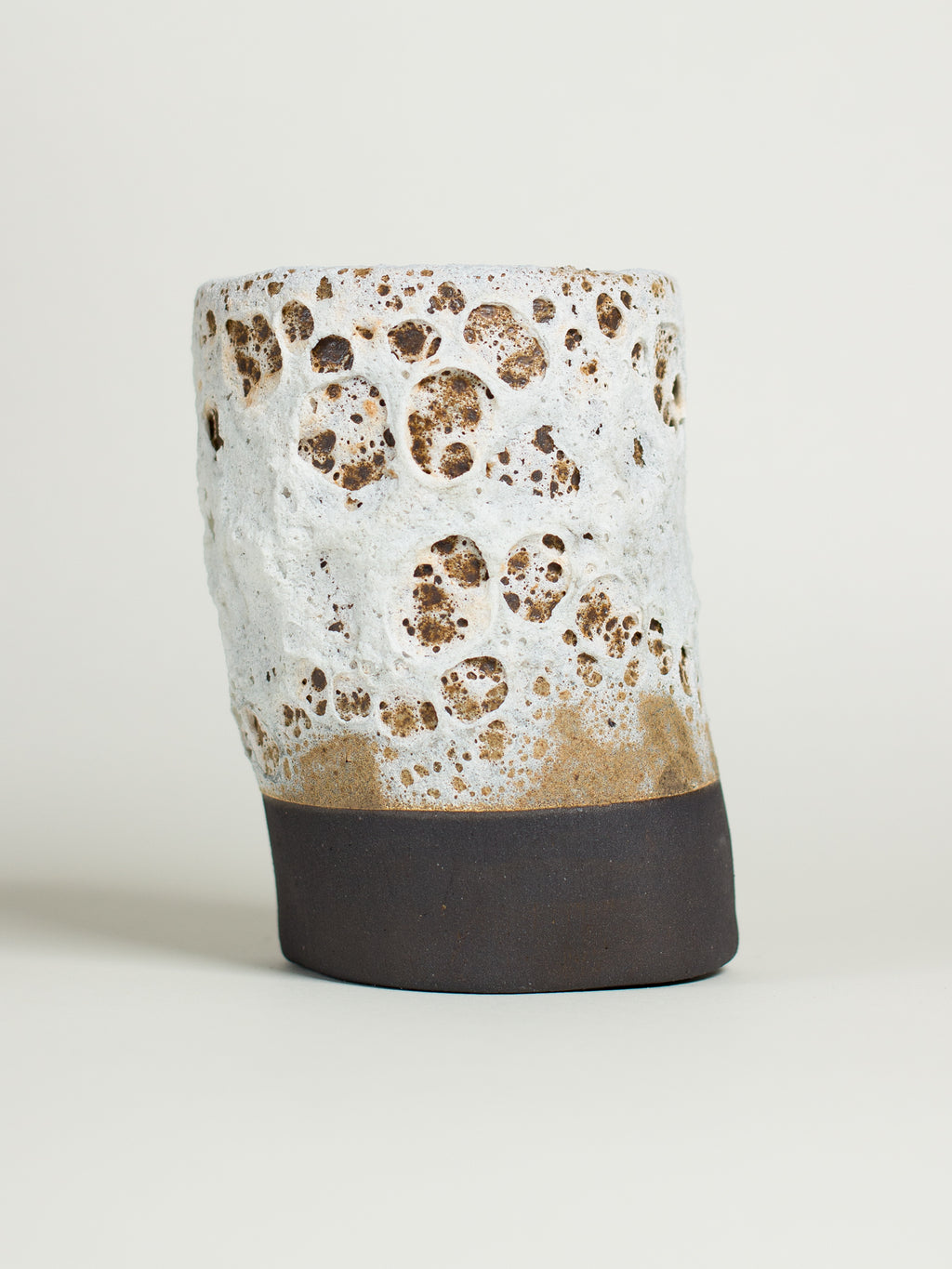 White Lava on a thrown cylinder of dark clay, white crust pitted with open brown craters