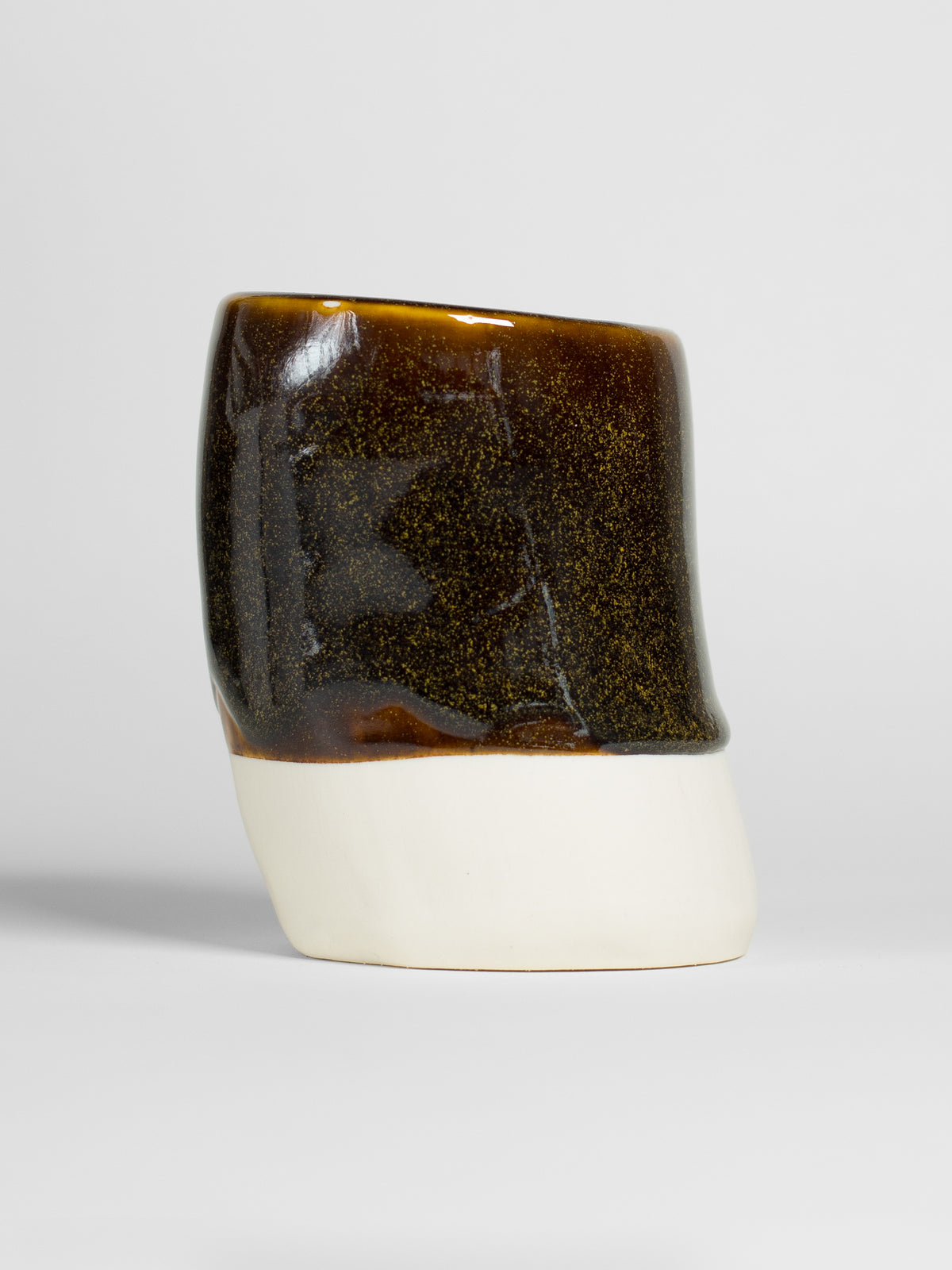 A handmade ceramic object with a tall, cylindrical shape. The upper portion is glazed with shiny, dark honey glaze that transforms into sparkly golden colour when thick. The cylinder features an artistic, organic design with a matte finish.