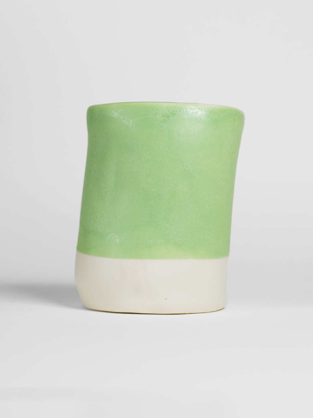A handmade ceramic object with a slightly tilted cylindrical shape. The upper half is glazed in a matt, satin salad green pottery glaze, while the bottom part is unglazed, revealing the natural white colour of the clay. The design is simple yet artistic with a smooth, polished finish.