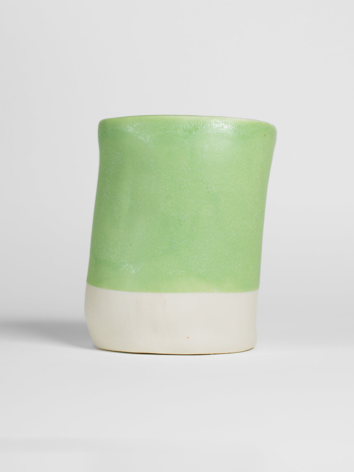 A handmade ceramic object with a slightly tilted cylindrical shape. The upper half is glazed in a matt, satin salad green pottery glaze, while the bottom part is unglazed, revealing the natural white colour of the clay. The design is simple yet artistic with a smooth, polished finish.