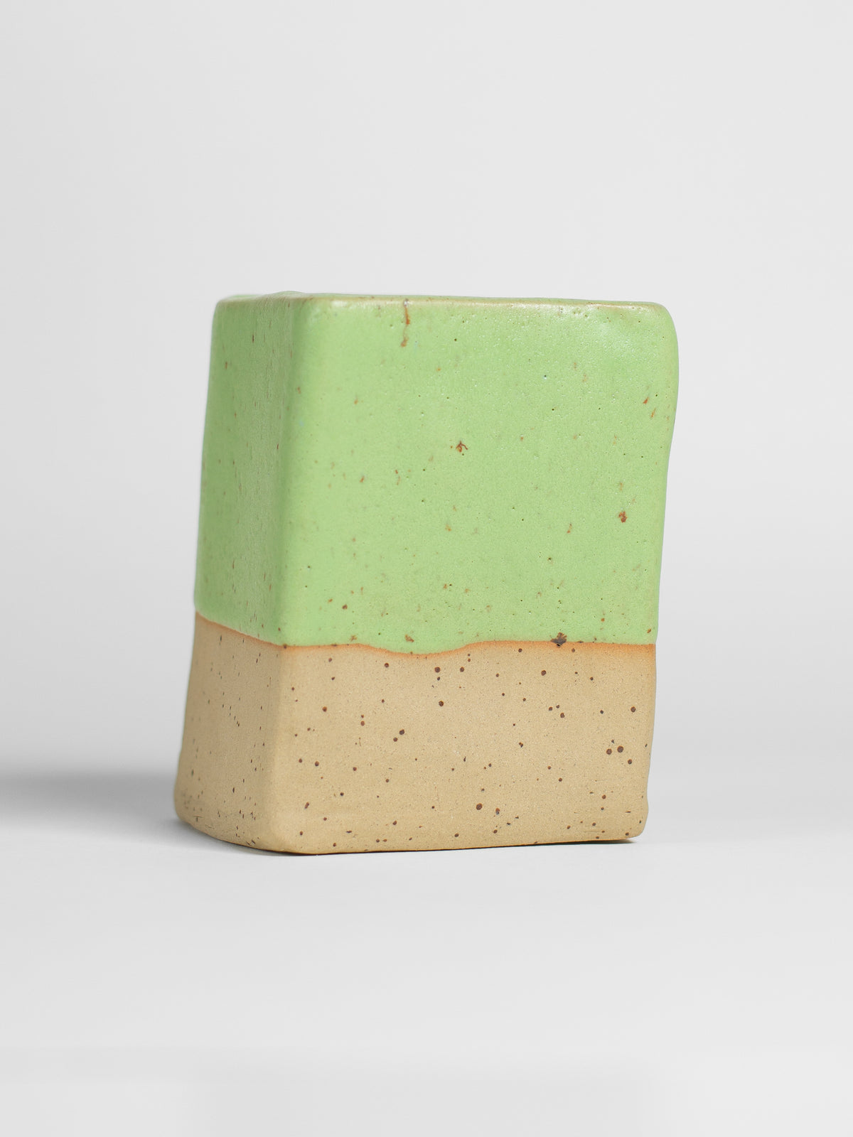 A handmade ceramic object with a slightly tilted cylindrical shape. The upper half is glazed in a matt, satin salad green pottery glaze, while the bottom part is unglazed, revealing the natural speckled and toastycolour of the clay. The design is simple yet artistic with a smooth, polished finish.