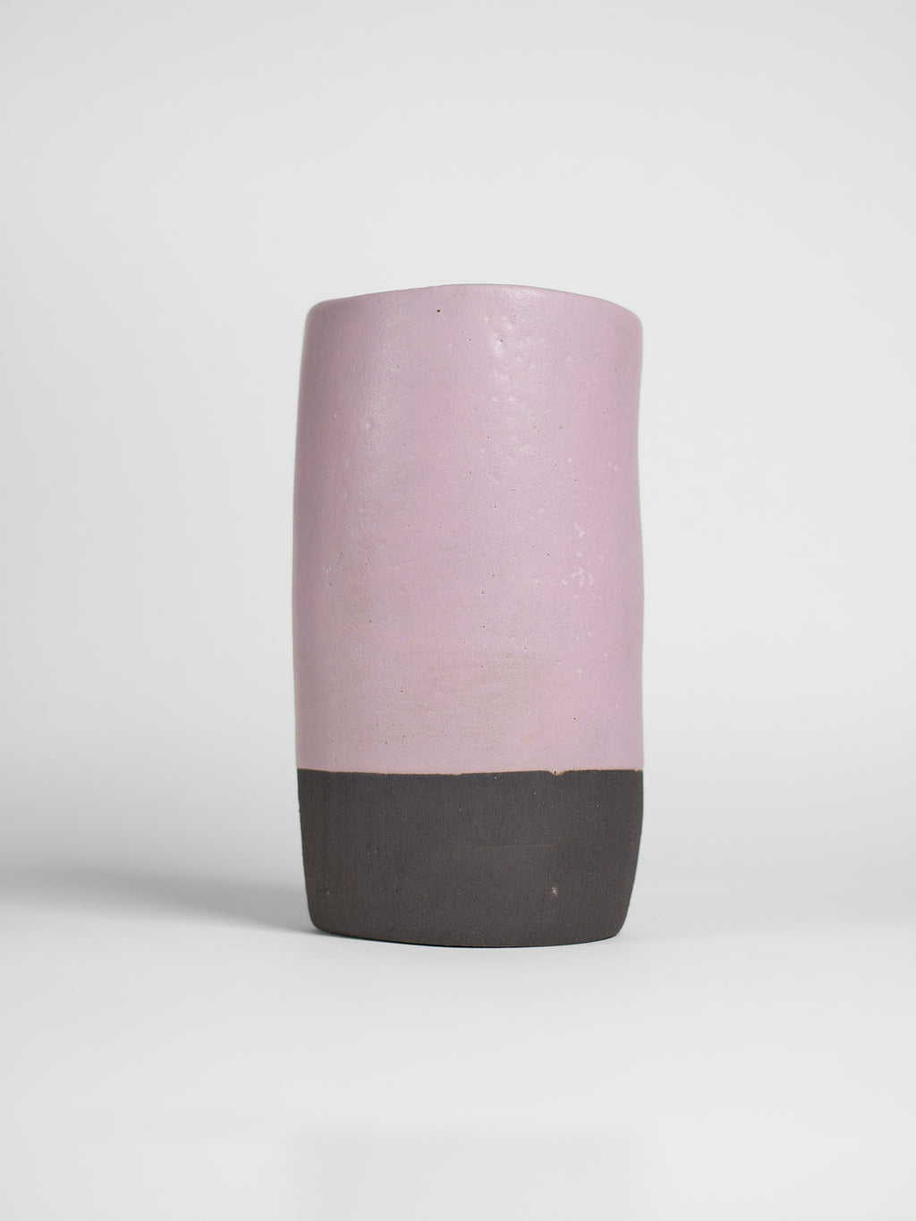 A handmade ceramic object with a slightly tilted cylindrical shape. The upper half is glazed in a matt, subtle violet , while the bottom part is unglazed, revealing the natural off-black colour of the clay. The design is simple yet artistic with a smooth, polished finish.