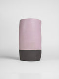 A handmade ceramic object with a slightly tilted cylindrical shape. The upper half is glazed in a matt, subtle violet , while the bottom part is unglazed, revealing the natural off-black colour of the clay. The design is simple yet artistic with a smooth, polished finish.