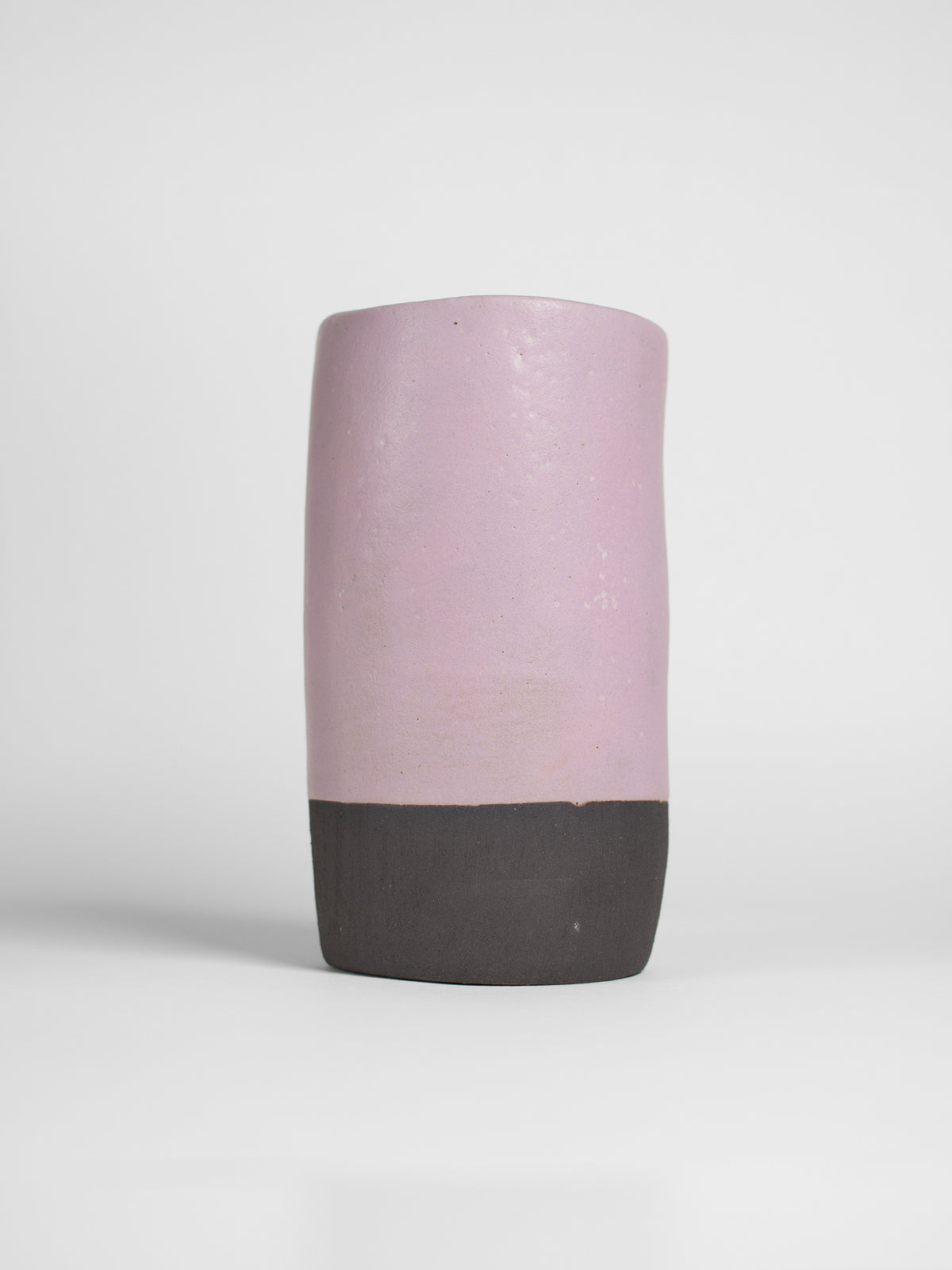 A handmade ceramic object with a slightly tilted cylindrical shape. The upper half is glazed in a matt, subtle violet , while the bottom part is unglazed, revealing the natural off-black colour of the clay. The design is simple yet artistic with a smooth, polished finish.