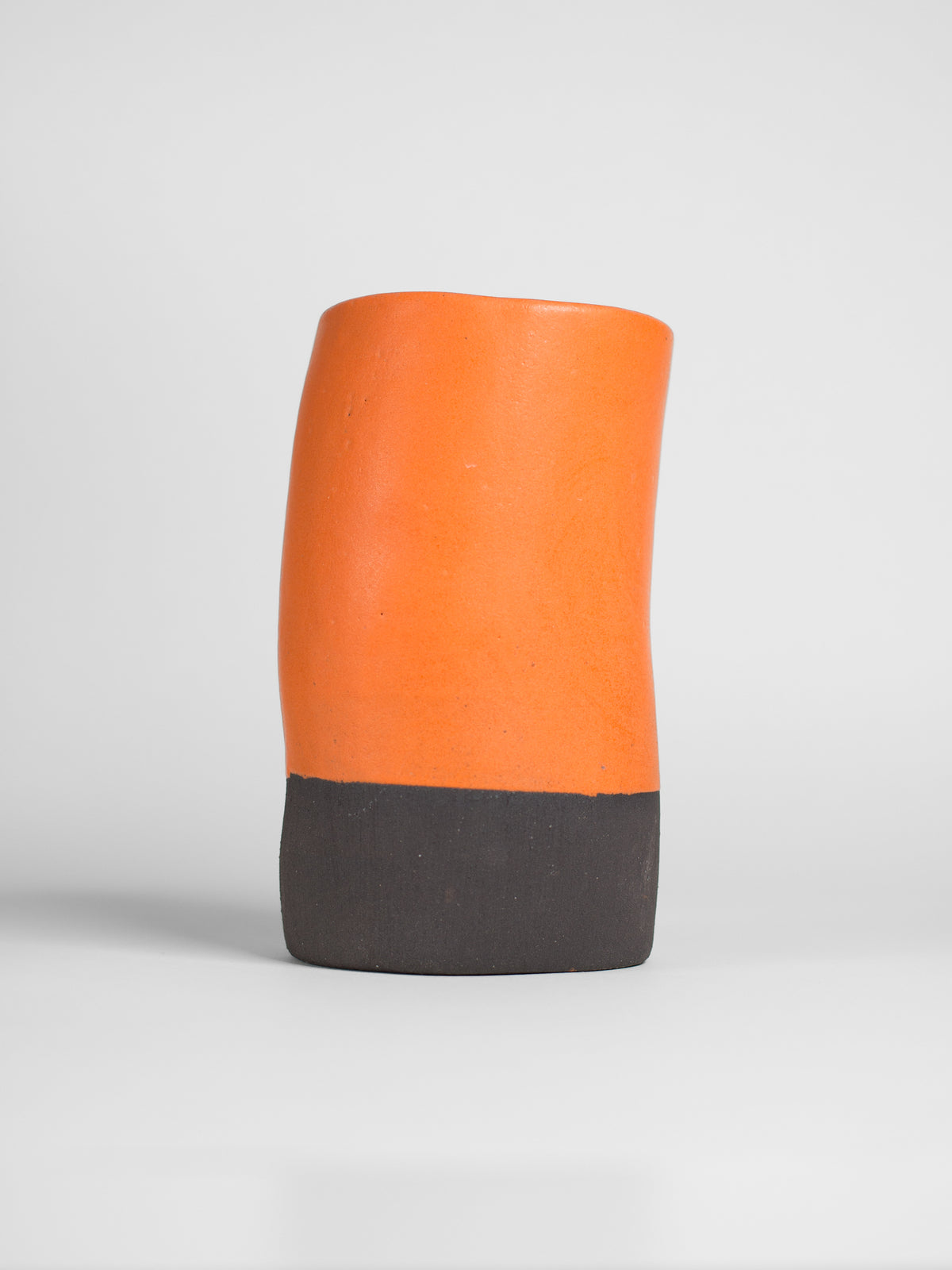 A handmade ceramic object with a slightly tilted cylindrical shape. The upper half is glazed in a matt, vibrant orange , while the bottom part is unglazed, revealing the natural off-black colour of the clay. The design is simple yet artistic with a smooth, polished finish.
