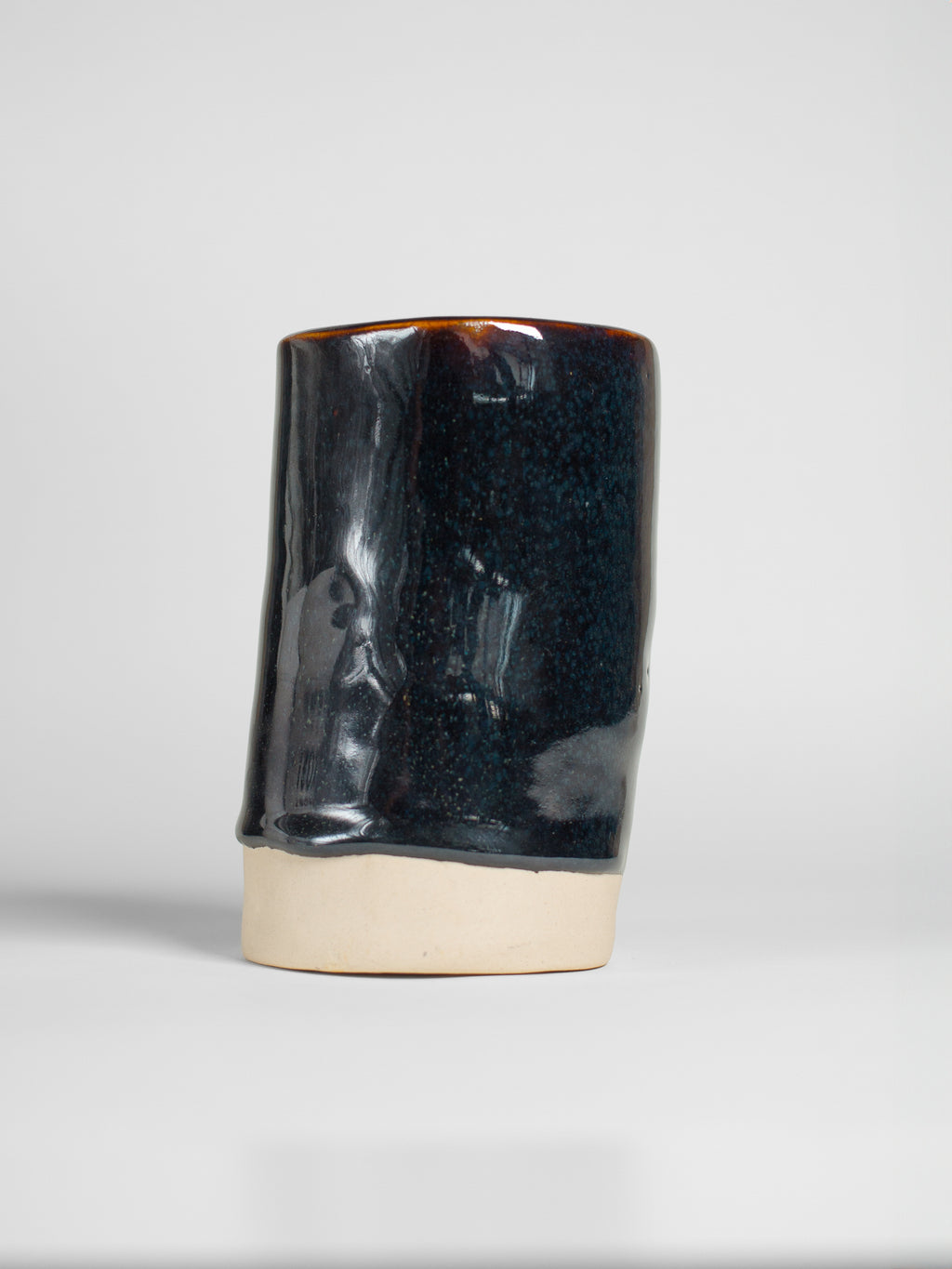 A handmade ceramic object with a tall, cylindrical shape. The upper portion is glazed in dark, shiny and reflective glaze with streaks. The cup features an artistic, organic design with a matte finish.