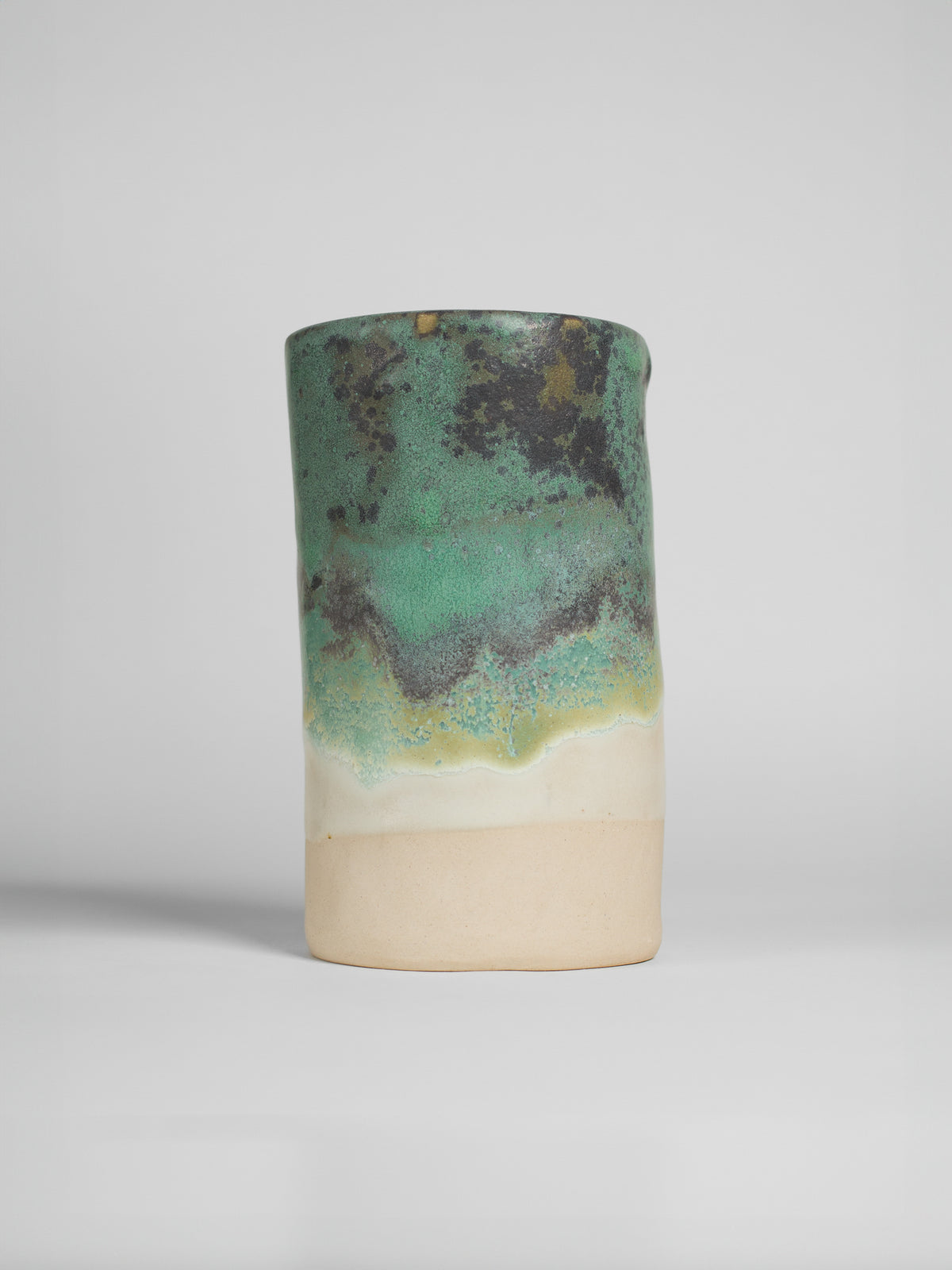 A handmade ceramic object with a tall, cylindrical shape. The upper portion is glazed in a textured green and black pattern, resembling a natural landscape, with hints of brown and gold. The middle section has soft green and yellow gradients, transitioning to the unglazed natural beige clay at the bottom. The cup features an artistic, organic design with a matte finish.