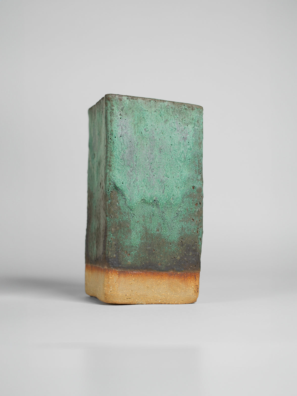 A handmade ceramic object with a tall, cylindrical shape. The upper portion is glazed in a textured green and black pattern, resembling a natural landscape, with hints of brown and gold. The middle section has soft green and yellow gradients, transitioning to the unglazed natural beige clay at the bottom. The cup features an artistic, organic design with a matte finish.