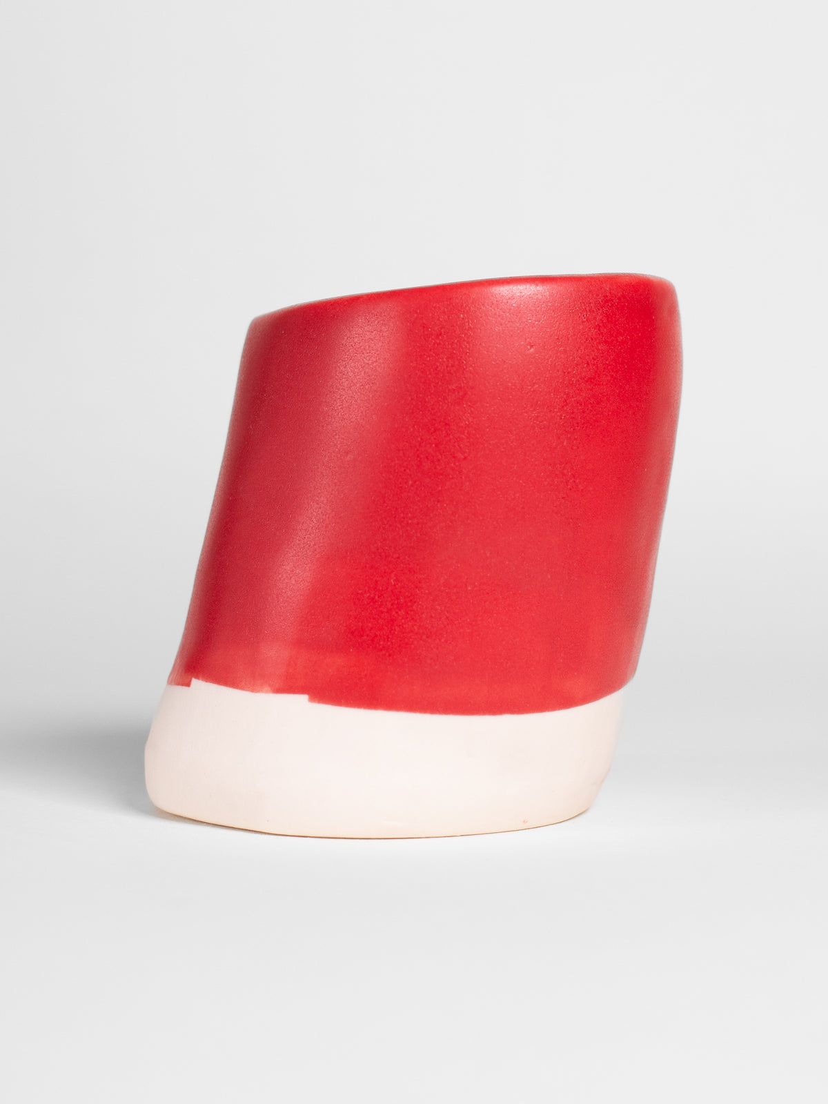 A handmade ceramic object with a slightly tilted cylindrical shape. The upper half is glazed in a matt, vibrant red pottery glaze, while the bottom part is unglazed, revealing the natural white colour of the clay. The design is simple yet artistic with a smooth, polished finish.