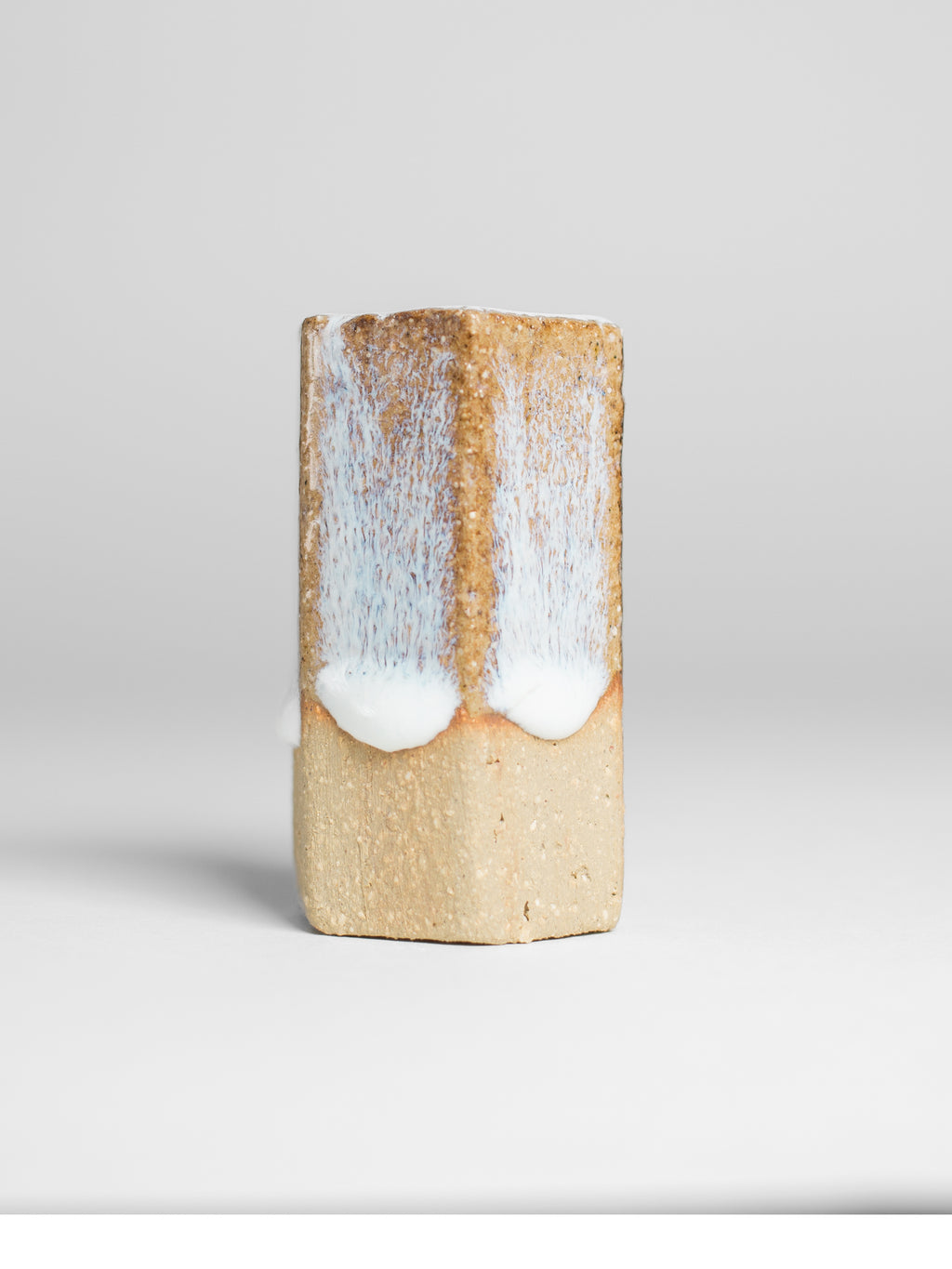 A handmade ceramic object with a tall, cylindrical shape. The upper portion is glazed in milky white and shiny glaze with off blue streaks. The cylinder features an artistic, organic design with a matte finish.