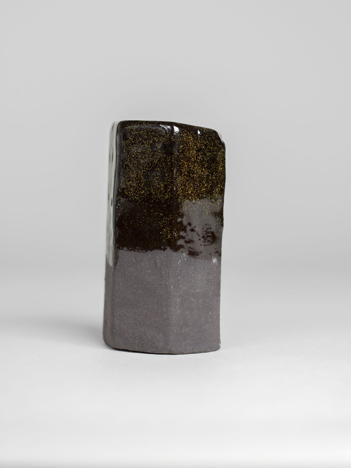 A handmade ceramic object with a tall, cylindrical shape. The upper portion is glazed with shiny, dark honey glaze that transforms into sparkly golden colour when thick. The cylinder features an artistic, organic design with a matte finish.