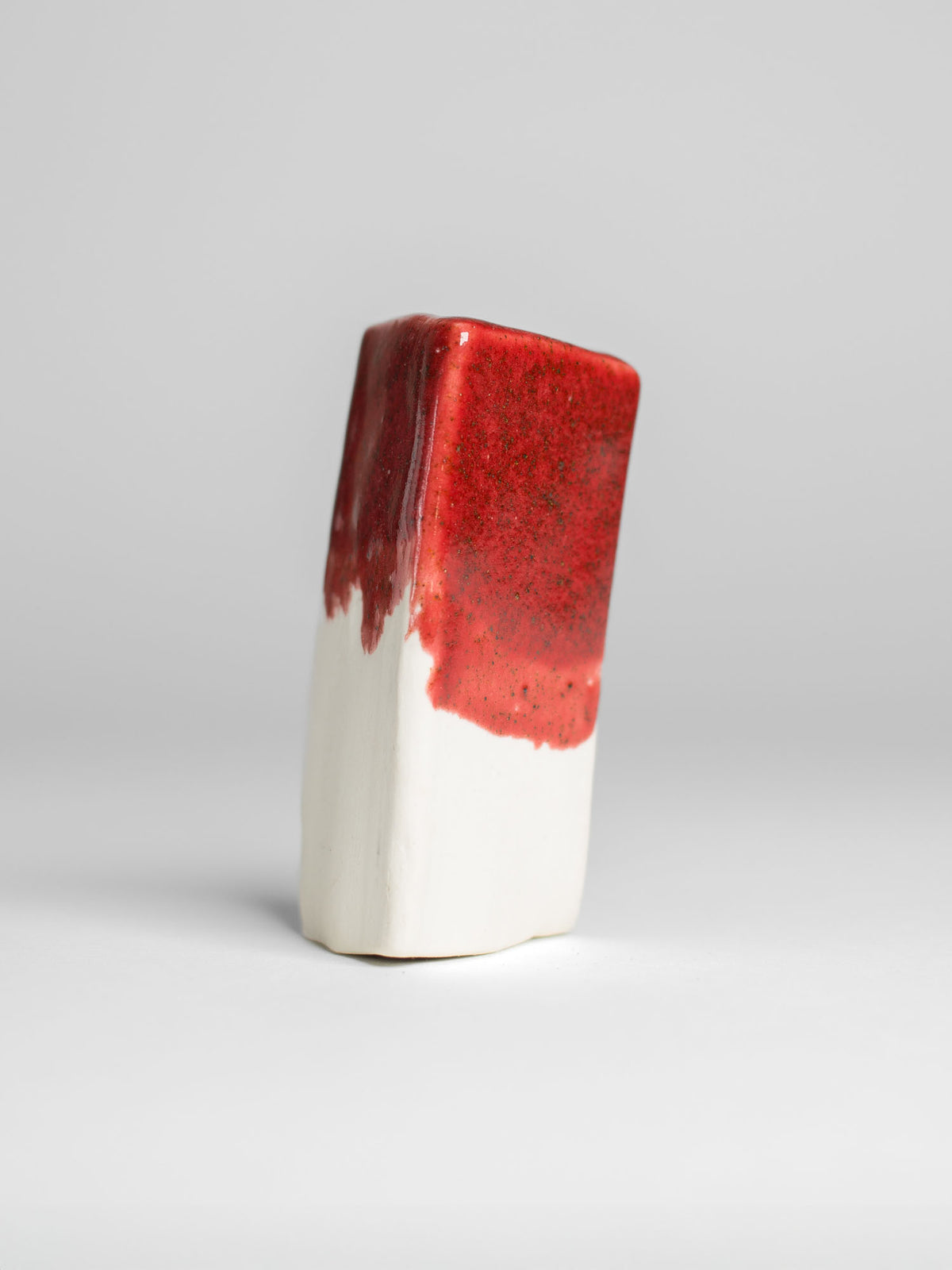 A handmade ceramic object with a tall, cylindrical shape. The upper portion is glazed in shiny red pottery glaze with golden speckle. The cup features an artistic, organic design with a matte finish.