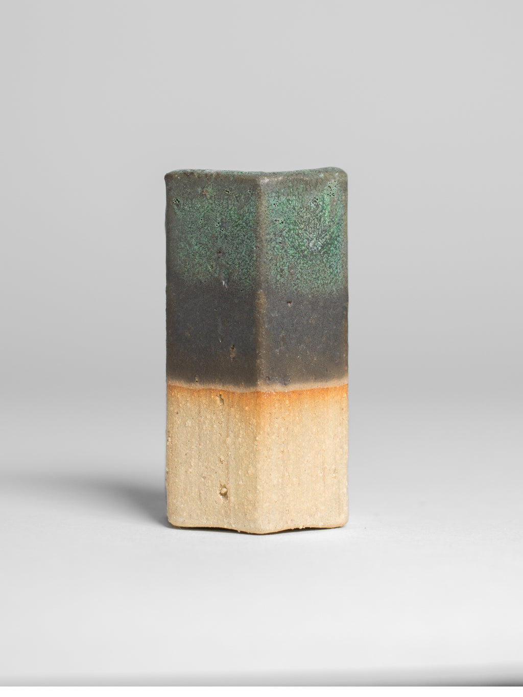 A handmade ceramic object with a tall, cylindrical shape. The upper portion is glazed in a textured green and black pattern, resembling a natural landscape, with hints of brown and gold. The middle section has soft green and yellow gradients, transitioning to the unglazed natural beige clay at the bottom. The cup features an artistic, organic design with a matte finish.