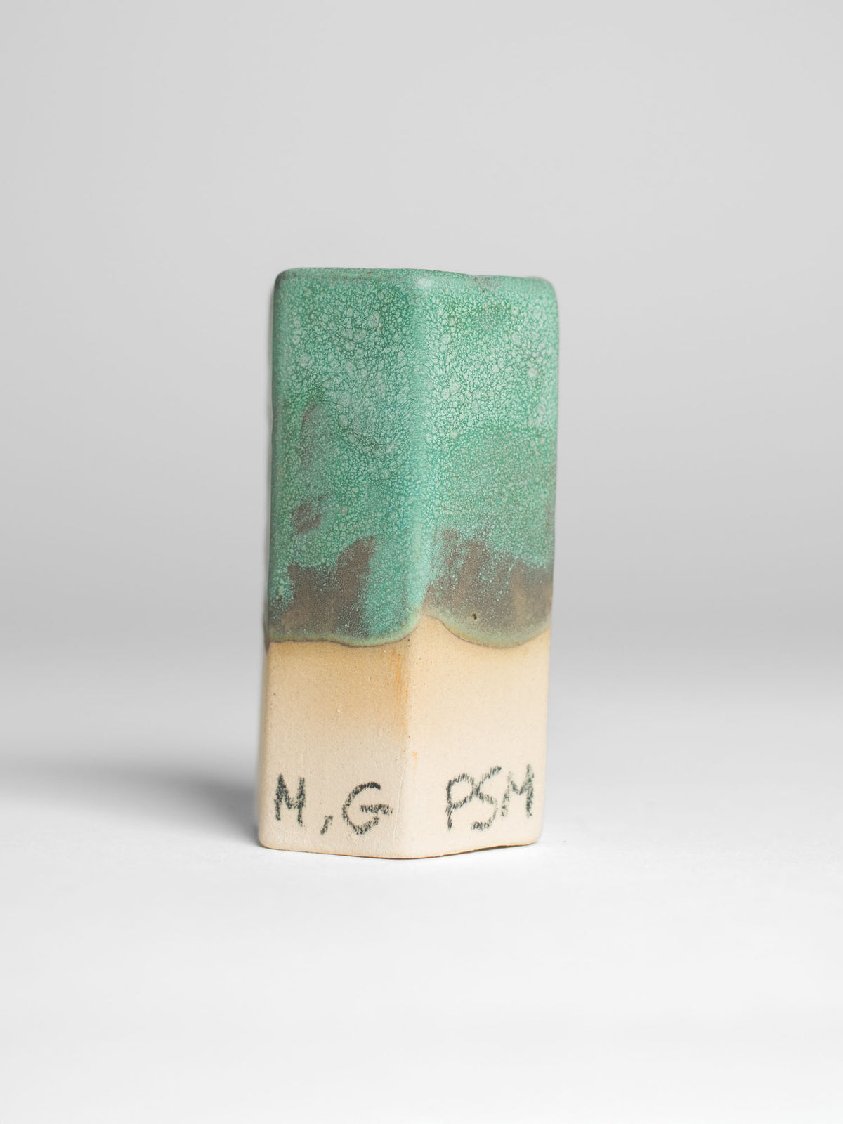 A handmade ceramic object with a tall, cylindrical shape. The upper portion is glazed in a textured green and black pattern, resembling a natural landscape, with hints of brown and gold. The middle section has soft green and yellow gradients, transitioning to the unglazed natural beige clay at the bottom. The cup features an artistic, organic design with a matte finish.