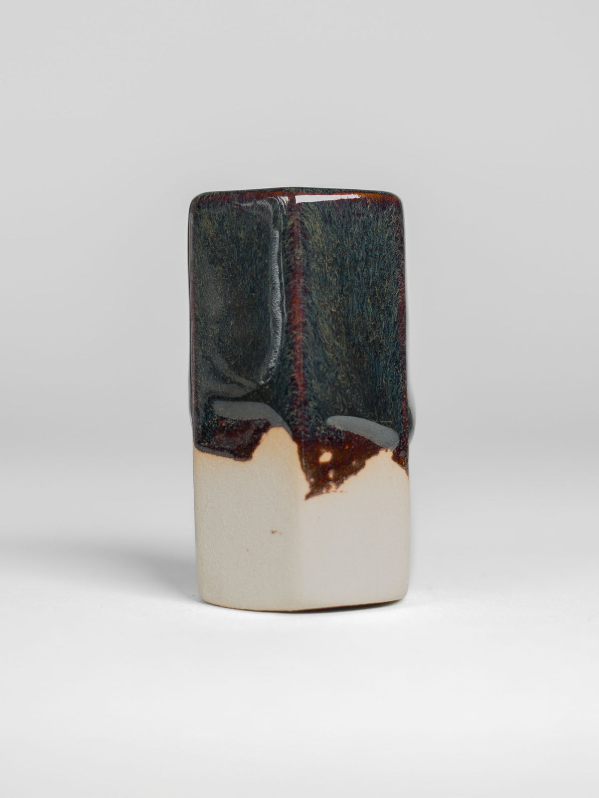 A handmade ceramic object with a tall, cylindrical shape. The upper portion is glazed in dark, shiny and reflective glaze with streaks. The cup features an artistic, organic design with a matte finish.