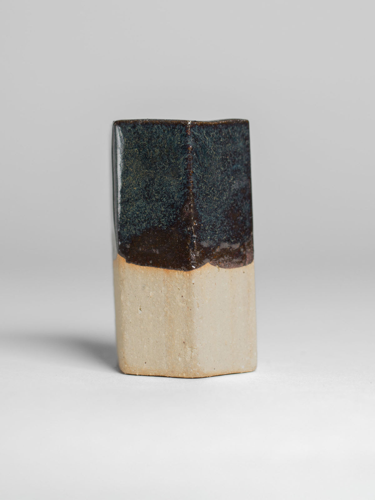 A handmade ceramic object with a tall, cylindrical shape. The upper portion is glazed in dark, shiny and reflective glaze with streaks. The cup features an artistic, organic design with a matte finish.