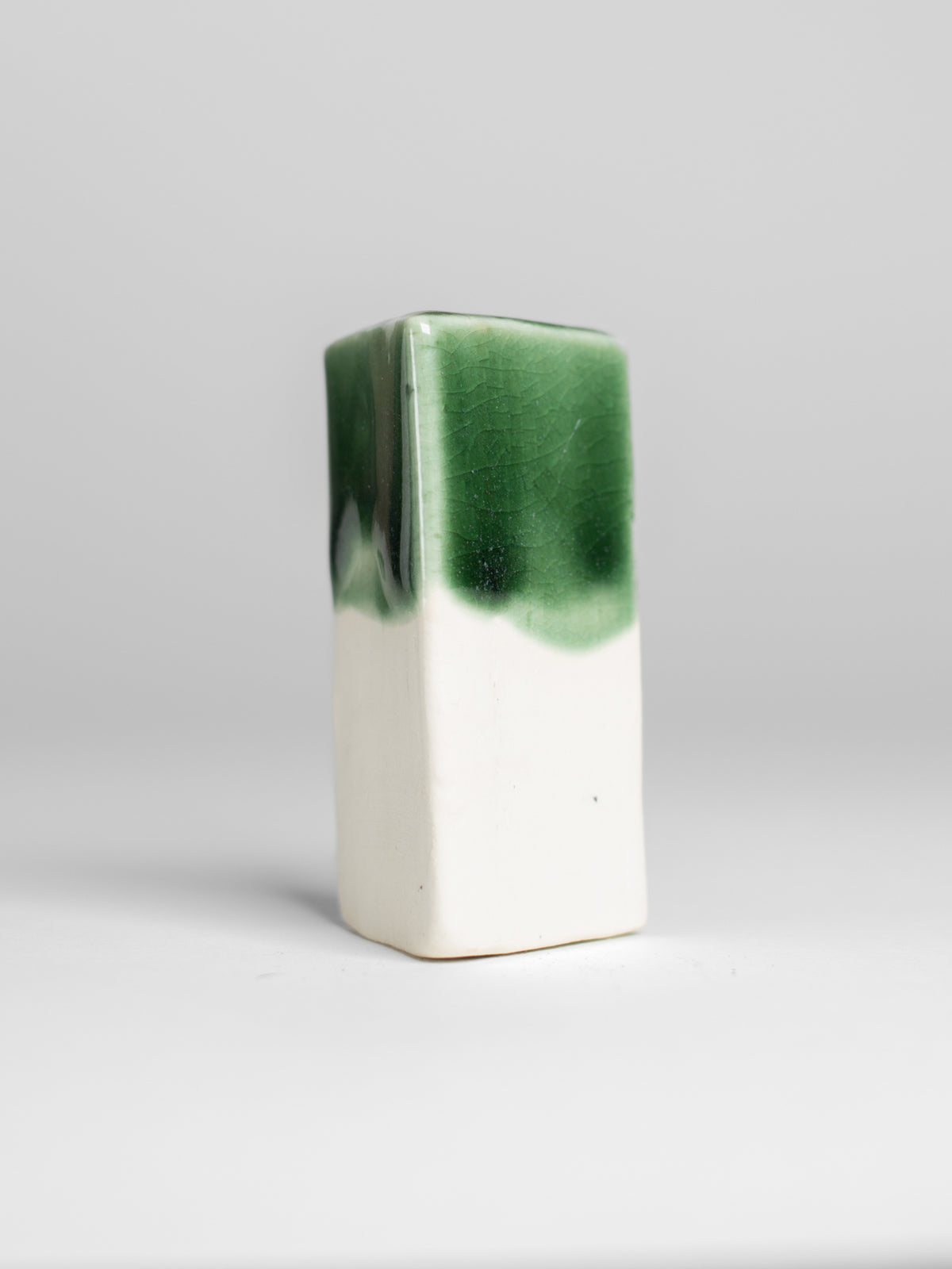 A handmade ceramic object. The upper half is glazed in a glossy, vibrant green with subtle variations in tone, while the bottom part is unglazed, revealing the natural off-white color of the clay. The design is simple yet artistic with a smooth, polished finish.