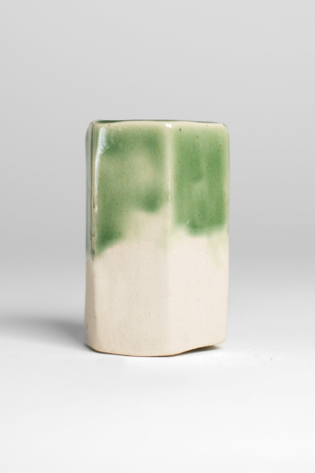 A handmade ceramic object with a slightly tilted cylindrical shape. The upper half is glazed in a glossy, vibrant green with subtle variations in tone, while the bottom part is unglazed, revealing the natural off-white coloyr of the clay. The design is simple yet artistic with a smooth, polished finish.