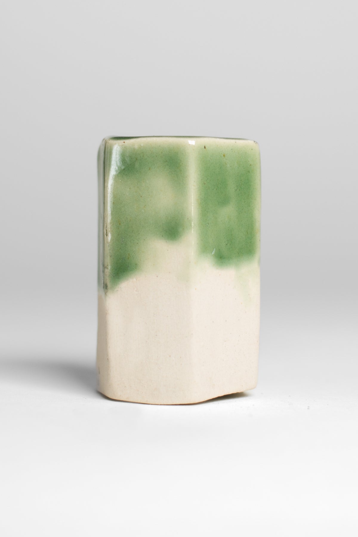 A handmade ceramic object with a slightly tilted cylindrical shape. The upper half is glazed in a glossy, vibrant green with subtle variations in tone, while the bottom part is unglazed, revealing the natural off-white coloyr of the clay. The design is simple yet artistic with a smooth, polished finish.
