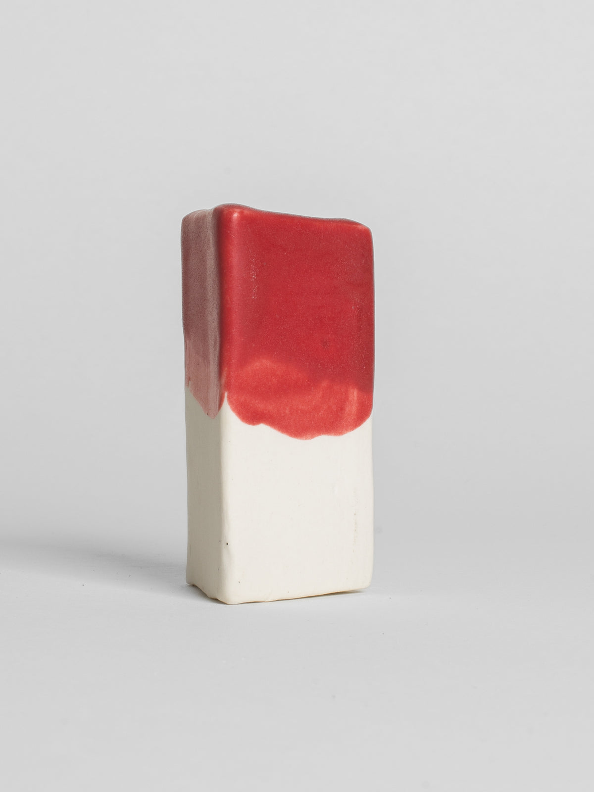 A handmade ceramic object with a slightly tilted cylindrical shape. The upper half is glazed in a matt, vibrant red pottery glaze, while the bottom part is unglazed, revealing the natural white colour of the clay. The design is simple yet artistic with a smooth, polished finish.