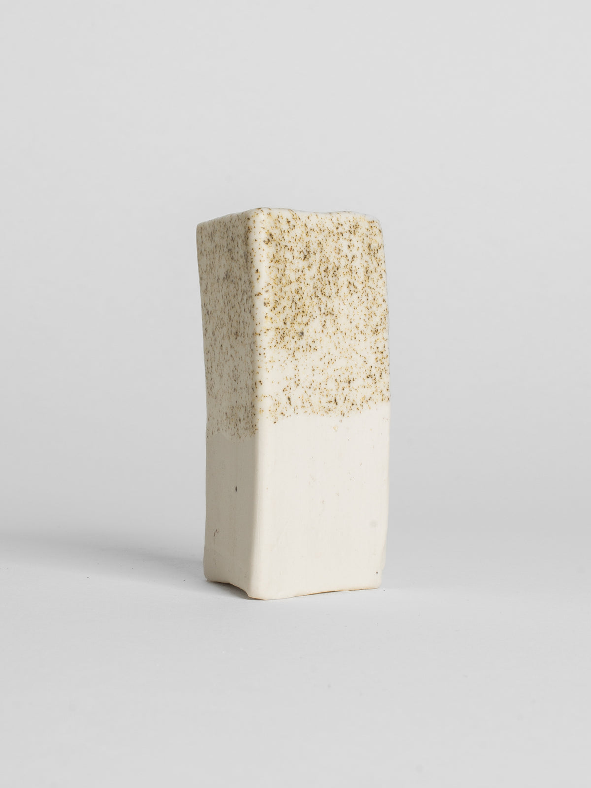 A handmade ceramic object with a tall, cylindrical shape. The upper portion is glazed in milky matt glaze with golden fleck. The cylinder features an artistic, organic design with a matte finish.