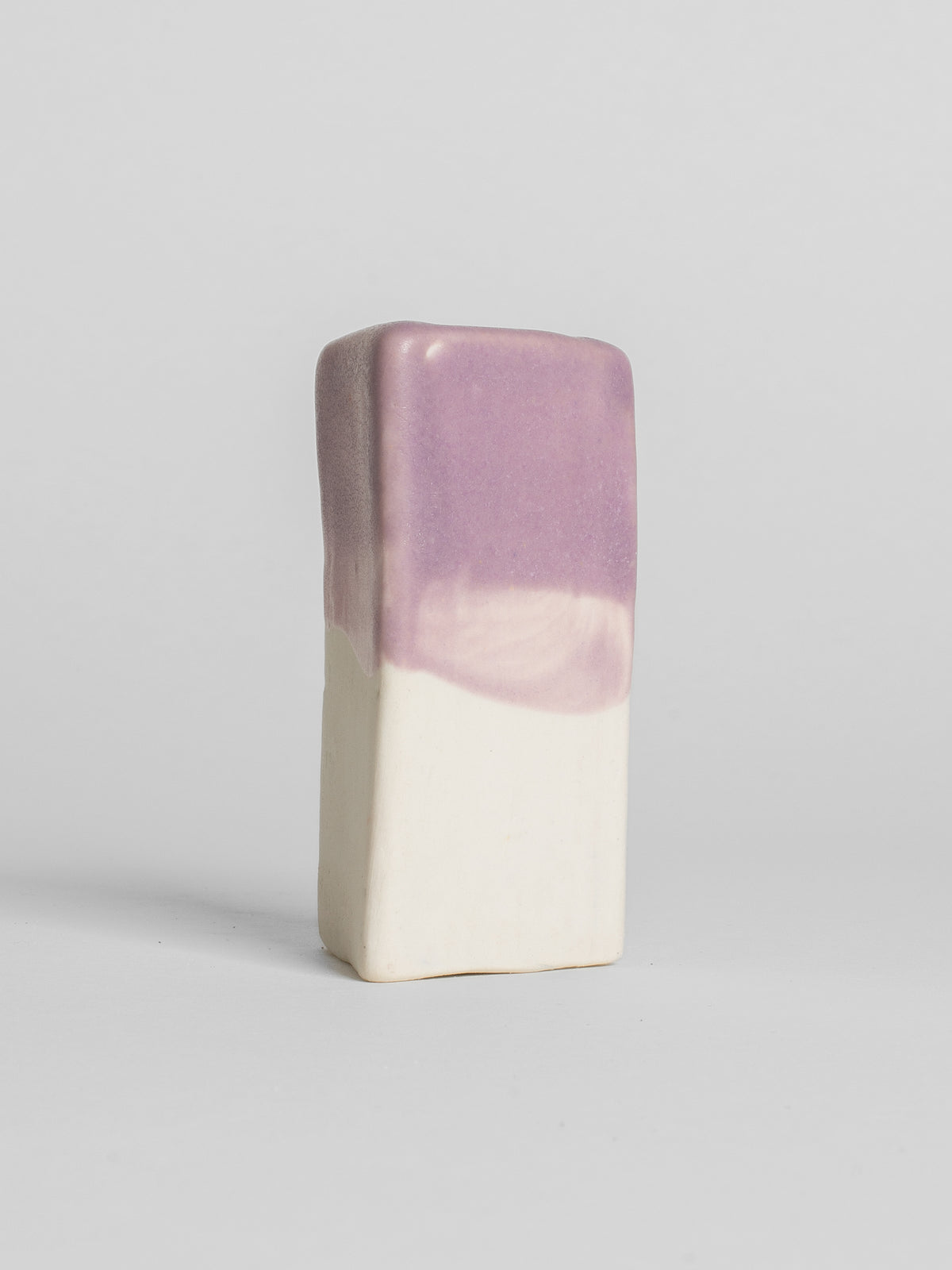 A handmade ceramic object with a slightly tilted cylindrical shape. The upper half is glazed in a matt, subtle violet , while the bottom part is unglazed, revealing the natural colour of the porcelain clay. The design is simple yet artistic with a smooth, polished finish.