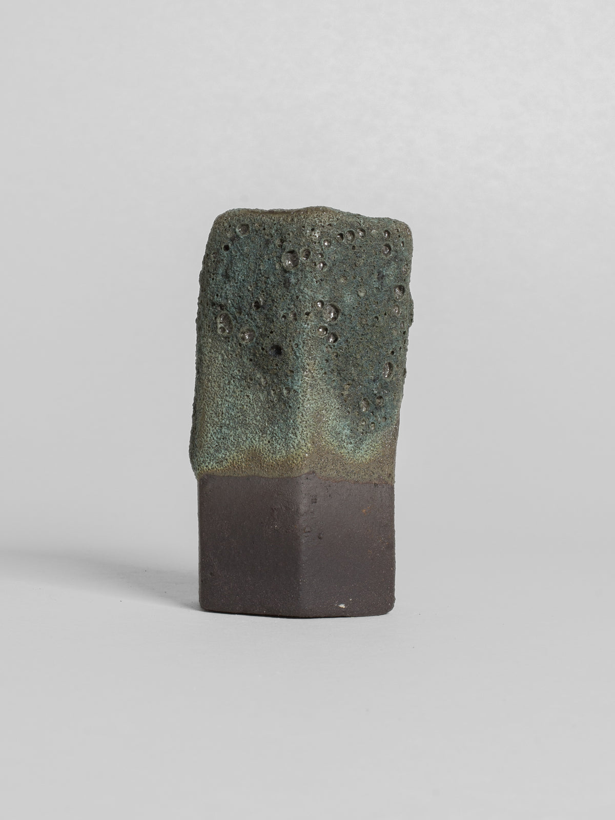 A handmade ceramic object with a tall, cylindrical shape. The upper portion is glazed in highly textured dark brown/bottle green lava glaze.