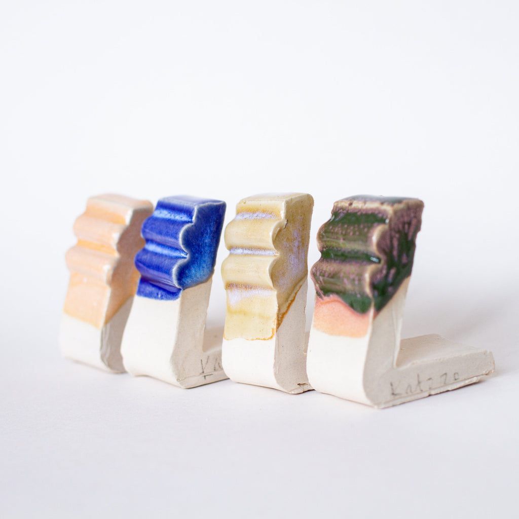 A row of four ceramic test tiles is displayed against a white background. Each tile features a different glaze color and finish, ranging from soft peach and vibrant blue to earthy yellow and dark brown-green. The test tiles have a grooved texture that shows how the glaze interacts with the surface, highlighting variation in colour and gloss. These tiles demonstrate the effects of different glaze formulations, useful for ceramic testing and experimentation.