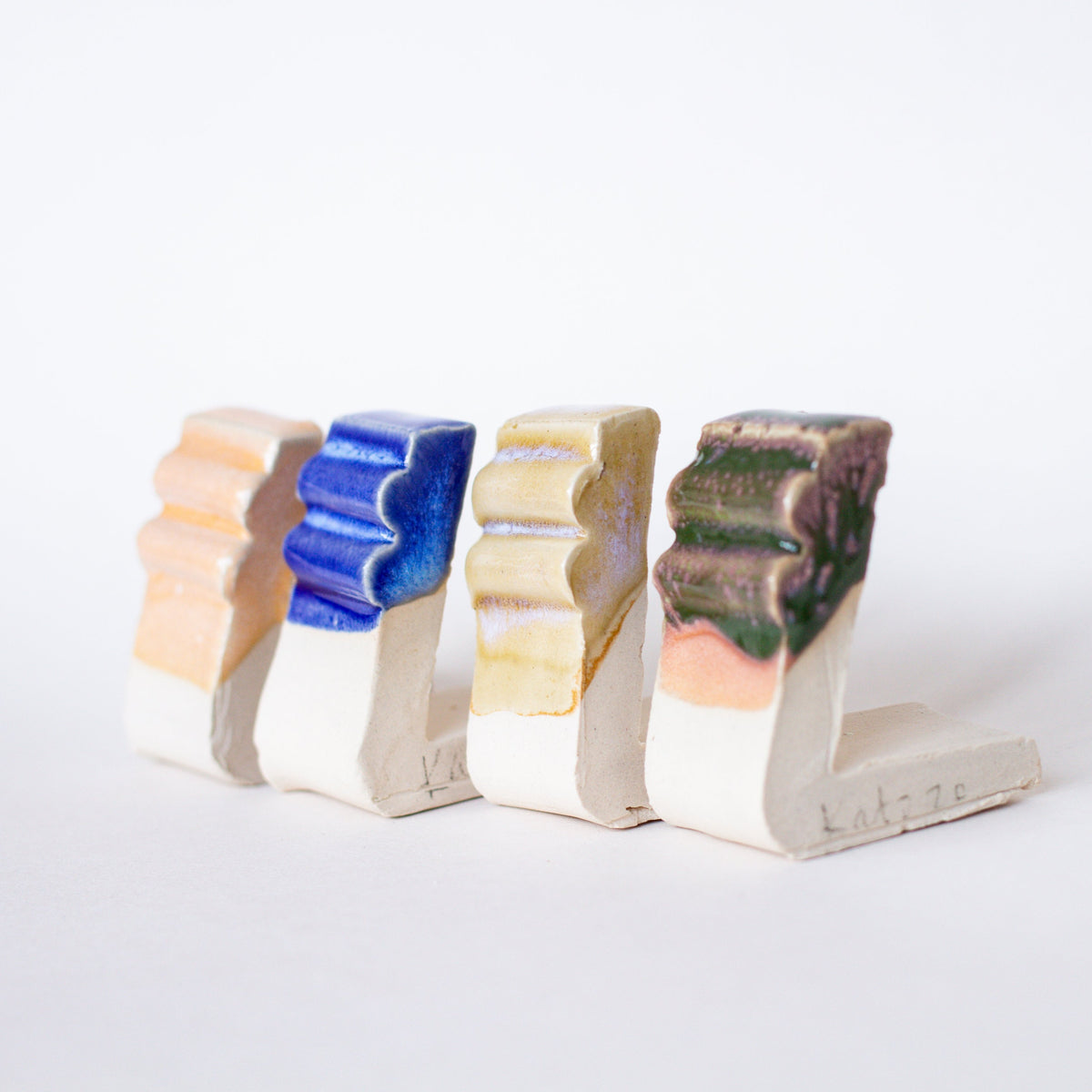 A row of four ceramic test tiles is displayed against a white background. Each tile features a different glaze color and finish, ranging from soft peach and vibrant blue to earthy yellow and dark brown-green. The test tiles have a grooved texture that shows how the glaze interacts with the surface, highlighting variation in colour and gloss. These tiles demonstrate the effects of different glaze formulations, useful for ceramic testing and experimentation.