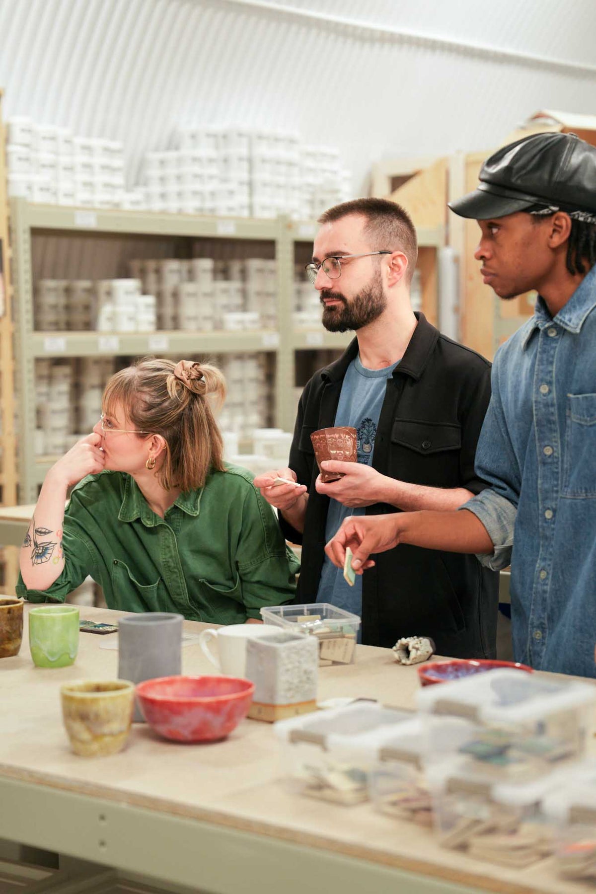 Three people in a pottery studio examining ceramic pieces on a table.