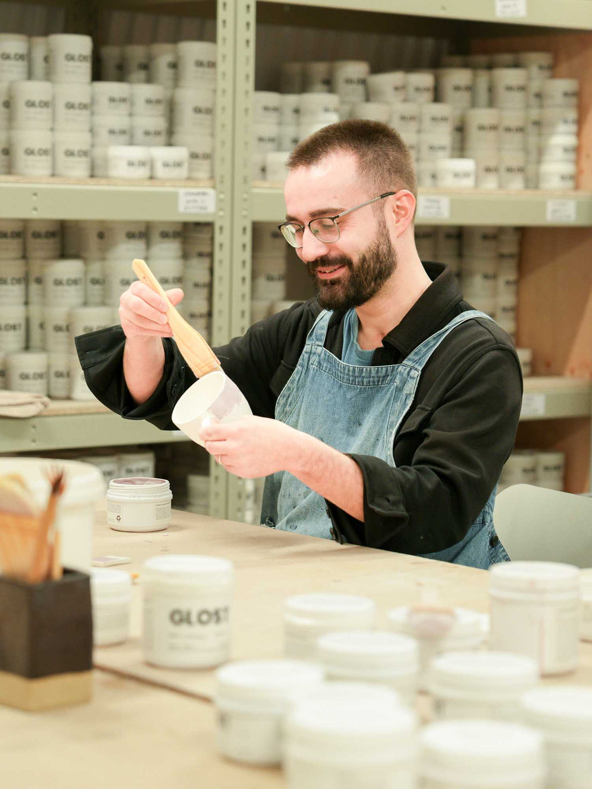 Man in apron holding a paintbrush in a pottery studio setting with shelves of paint cans.