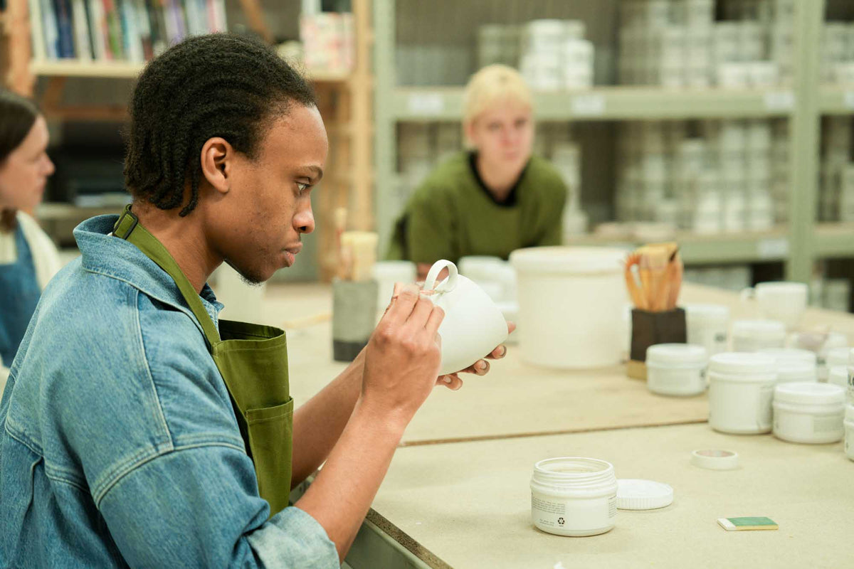 Person holding a ceramic cup in a pottery studio with shelves of clay pots in the background.