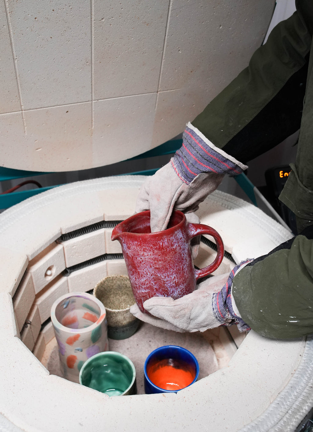 A gloved hand lifting a red mug from an electric kiln during unloading.