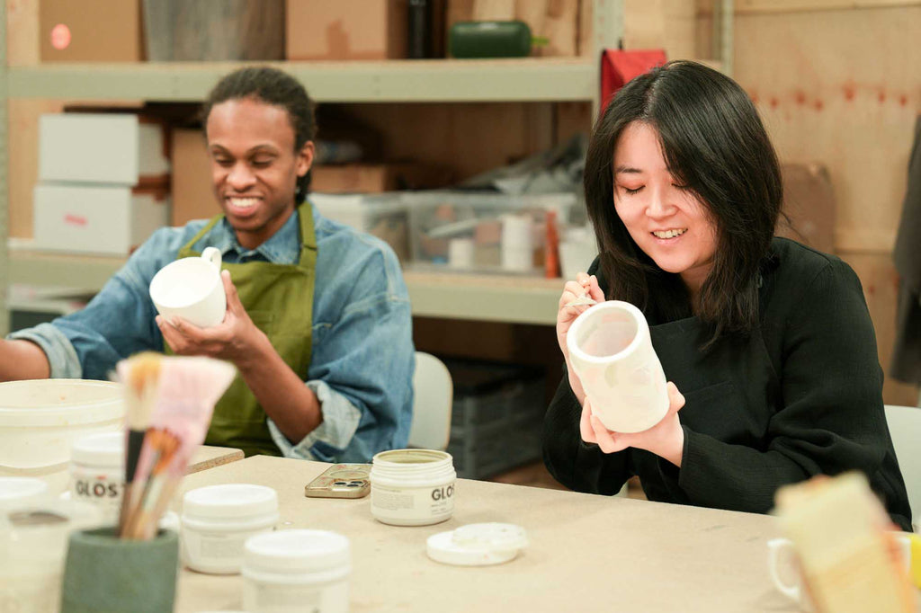Two people in a pottery studio holding ceramic pieces and brushing on glaze 