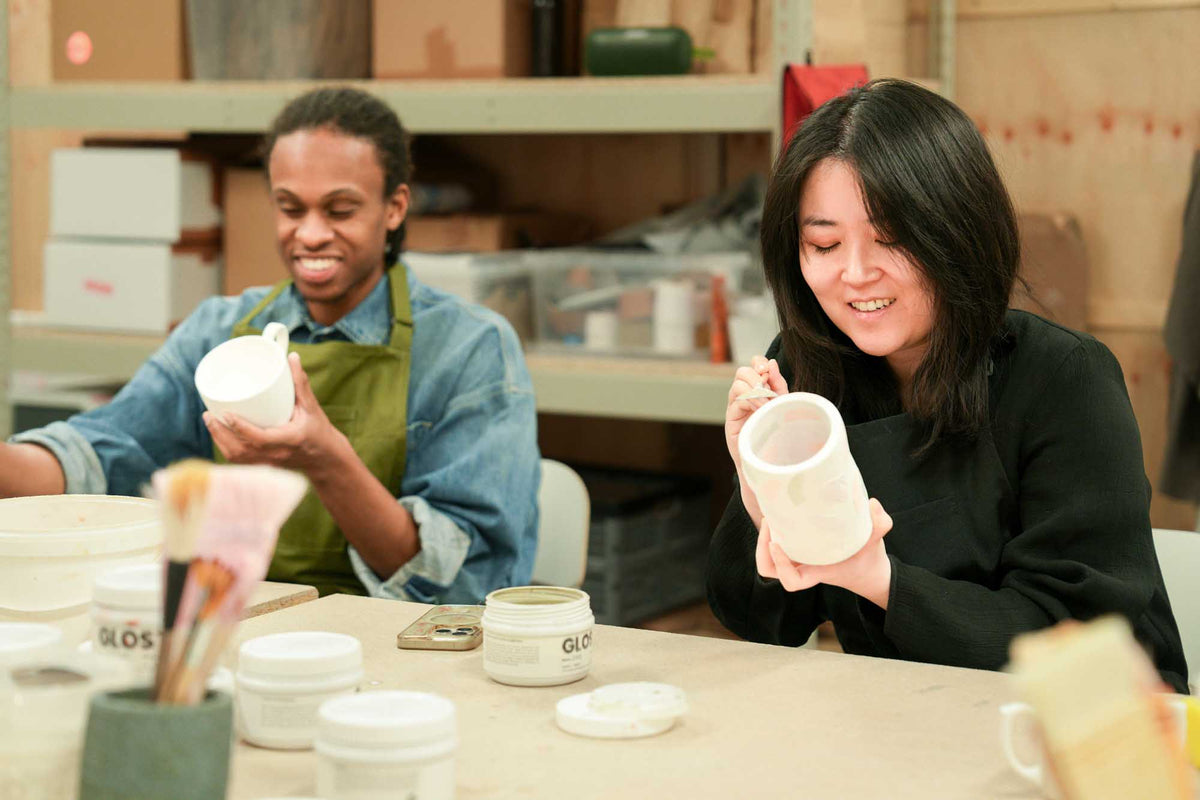 Two people in a pottery studio holding ceramic pieces and brushing on glaze 