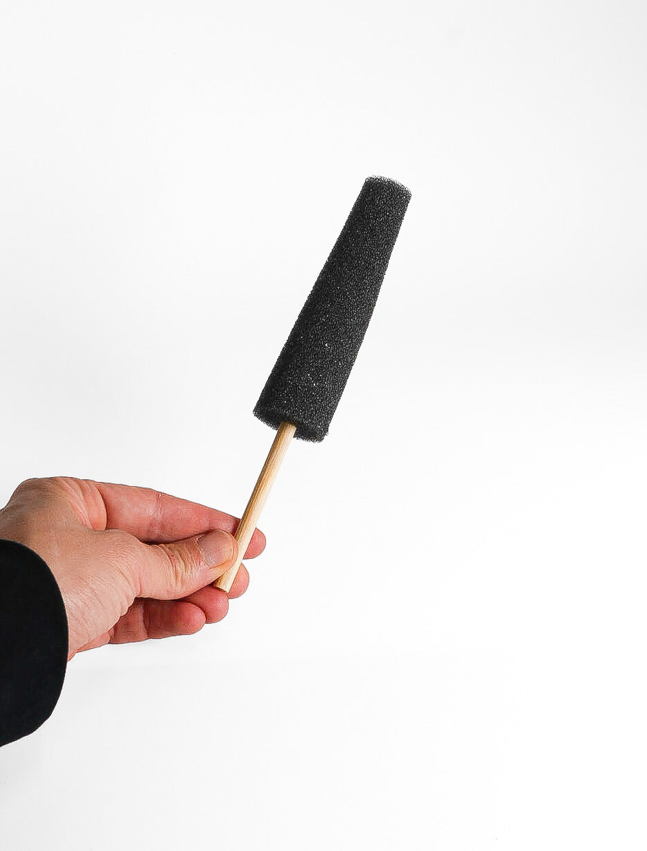 A black tapered foam sponge on a slim wooden stick, held in one hand against a white background.