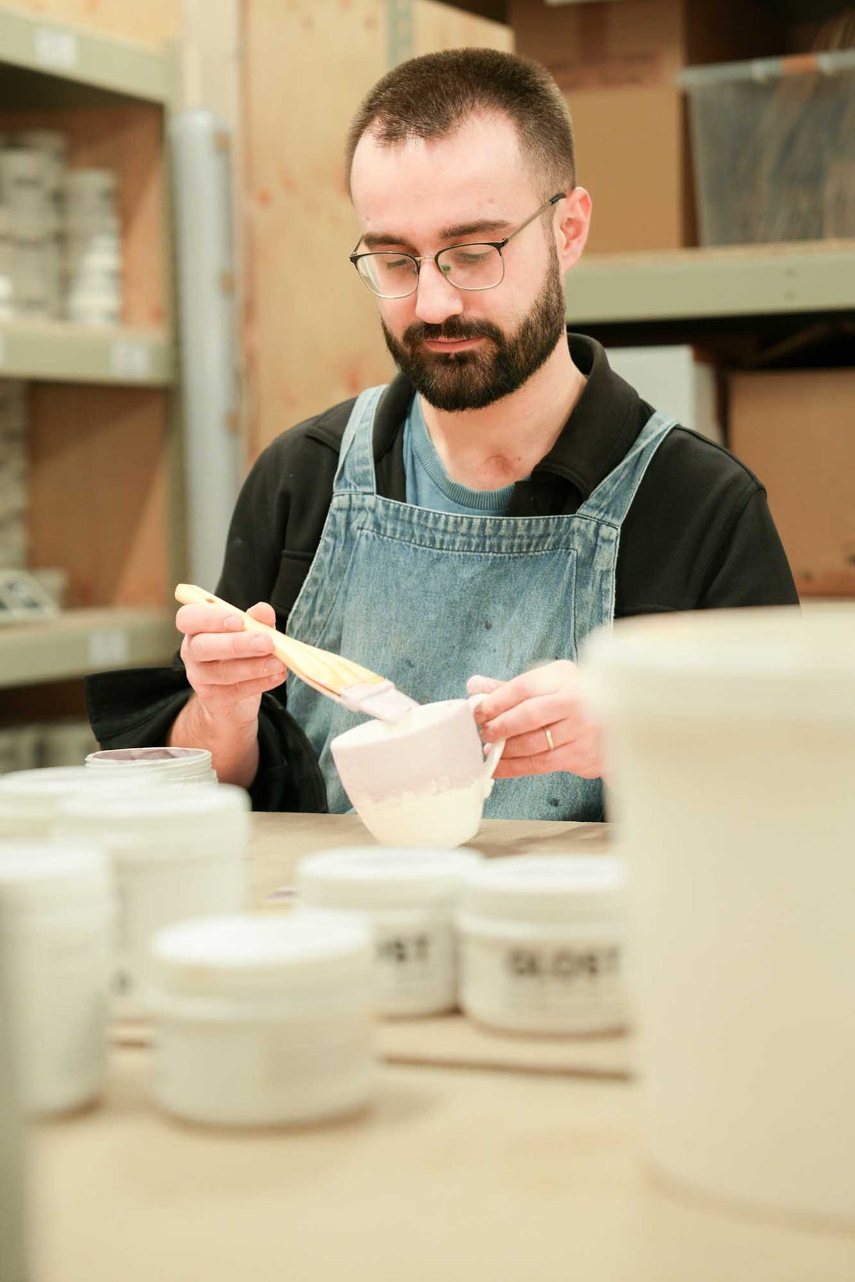 A maker in a denim apron brushing white glaze onto a small ceramic jug, with GLOST glaze jars on the table
